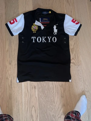 Ralph Lauren Tokyo Polo - Säljer en Polo Ralph Lauren pikétröja i helt nytt skick – aldrig använd. Tröjan är i svart med vita detaljer och exklusiva broderier, inklusive TOKYO-text, klassisk Polo-logga och detaljer på ärmarna som ger ett sportigt och unikt uttryck.  Inga skador, fläckar eller slitage – i perfekt skick.  En stilren och iögonfallande piké av hög kvalitet, perfekt till både vardag och finare tillfällen.  Hör av dig vid frågor eller om du vill ha fler bilder.