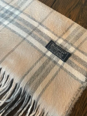 Beige rutig halsduk från Burberrys - Klassisk beige halsduk från Burberrys i 100% kashmir. Den har det ikoniska rutmönstret i beige, grått och vitt samt fransar i kanterna. Perfekt accessoar för att lyfta din outfit med en tidlös och lyxig känsla.