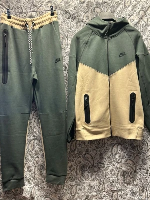 Nike tvådelad tracksuit  set grön/beige - Nike tracksuit set med hoodie och byxor i grönt och beige. Jackan har dragkedja, huva och svarta detaljerade fickor. Byxorna har resår i midjan med snörning och matchande svarta dragkedjor. Perfekt för chill dagar hemma eller på språng.