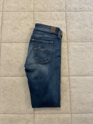 Blå slim fit jeans från Replay - Snygga blå slim fit jeans från Replay med klassisk femficksdesign och slitningar på låren. Jeansen har en mörk tvätt med ljusare partier och kontrastsömmar. Perfekta för en trendig och avslappnad stil.