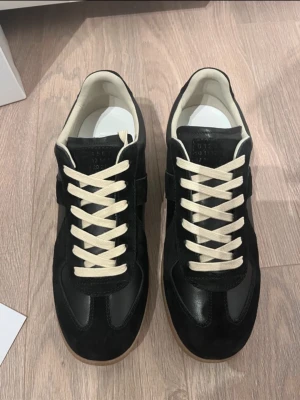 Svarta Maison Margiela gats  - Maison Margiela Replica sneakers i svart mocka och läder med vita kontrasterande skosnören och vit insida. Klassisk låg modell med retrovibbar och diskret logotyp på plösen. Snygg och tidlös design som passar till det mesta.