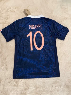Frankrike fotbollströja Mbappé #10 - Snygg blå fotbollströja från Nike med Frankrikes landslagsemblem och nummer 10 Mbappé på ryggen. Tröjan har vita och röda detaljer, vit krage och är tillverkad i ett lätt, ventilerande material perfekt för träning eller match.