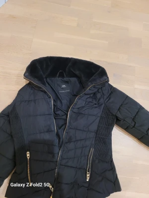 Svart pufferjacka från Zara - Snygg svart pufferjacka från Zara med mjuk fuskpäls i kragen och guldfärgade dragkedjor. Jackan har quiltad design, ribbade paneler på sidorna och två fickor med dragkedja. Perfekt för kalla dagar och har en modern, stilren look.