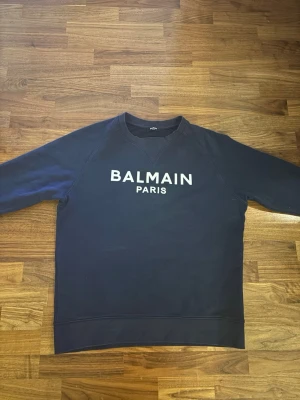 Gammal model Mörkblå sweatshirt från Balmain - Gammal model Snygg mörkblå sweatshirt från Balmain Paris med vit logga tryckt på bröstet. Klassisk rund halsringning och ribbade muddar vid ärmslut och nederkant. Perfekt för en trendig och avslappnad stil.