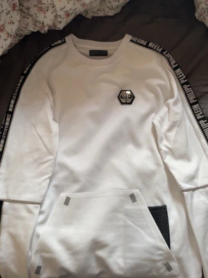 Vit Philipp Plein hoodie med logga - Snygg vit hoodie från Philipp Plein med svart logga på bröstet och svarta band med text längs ärmarna. Stor ficka framtill med coola detaljer och mesh på ena sidan. Tillverkad i mjukt material, perfekt för en streetwear-look.