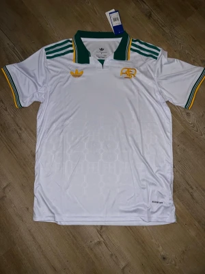 Vit roma fotbollströja Adidas - Snygg vit retroinspirerad fotbollströja från Adidas med gröna och gula detaljer. Tröjan har grönt krage, gula och gröna ränder på axlar och ärmslut samt broderade loggor på bröstet. Tillverkad i lätt och ventilerande material med Aeroready-teknologi.