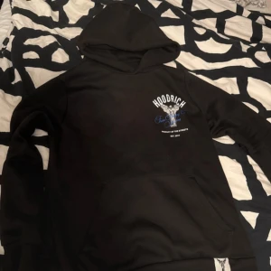 Svart HOODRICH hoodie med ängeltryck - Svart hoodie från HOODRICH med stor ängel och texten 'From Nothing To Something' i blått och vitt på ryggen. Liten logga och motiv på bröstet. Klassisk känguruficka och huva. Perfekt streetwear-vibe och riktigt snygg detalj på ryggen.