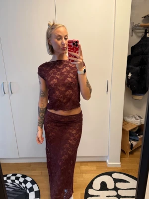 Vinrött spetsset från Gina Tricot - Tvådelat set i vinröd spets från Gina Tricot. Setet består av en kortärmad topp och en lång kjol, båda i genombruten spets med blommönster. Materialet är stretchigt och följsamt, perfekt för en trendig look.