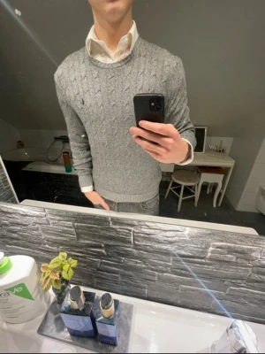 Grå kabelstickad tröja Polo Ralph Lauren - Säljer en grå kabelstickad tröja från Polo Ralph Lauren. Tröjan har rund halsringning, ribbade muddar och diskret logga på bröstet. Perfekt att bära över skjorta för en clean och klassisk look.