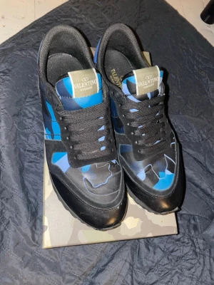 Valentino blå och svarta sneakers - Snygga sneakers från Valentino med svart och blå kamouflagemönster, svarta skosnören och detaljer i mocka och läder. Tungan har en guldfärgad logotyp. Perfekta för dig som vill sticka ut med exklusiva skor.