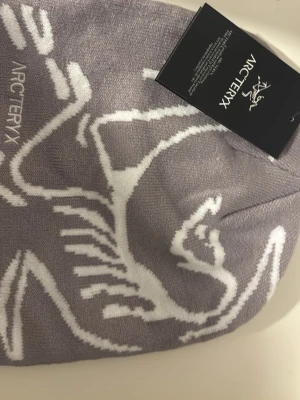Ljusgrå mössa från Arc'teryx - Snygg ljuslila mössa från Arc'teryx med vit logga och grafiskt mönster. Tillverkad i mjukt, stickat material som sitter skönt på huvudet. Perfekt accessoar för att hålla värmen och addera stil till din outfit.