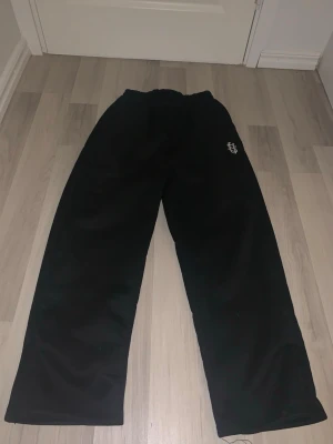 Black baggy joggers  - Svarta mjukisbyxor med vit broderad logga på benet. Byxorna har en avslappnad passform och resår i midjan. Perfekta för chill dagar eller när du vill vara bekväm. Materialet känns mjukt och skönt mot huden.