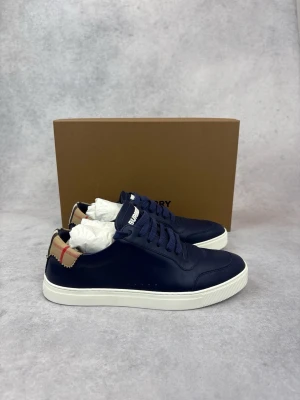 Marinblå Burberry sneakers - Snygga marinblå sneakers från Burberry med klassiskt rutigt tygdetalj vid hälen och vit sula. Skorna har rund tå, matchande blå skosnören och BURBERRY-logga på plösen. Tillverkade i slätt läder för en clean och stilren look.
