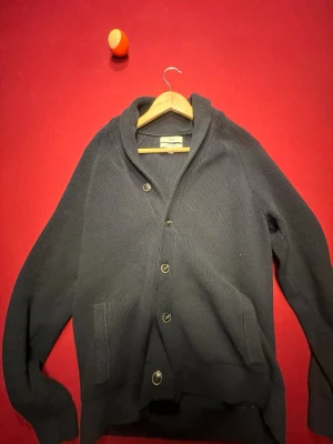 Mörkblå kofta från M&S - Snygg mörkblå kofta från M&S i bomullsblandning. Koftan har sjalkrage, knäppning med fyra knappar framtill och två fickor. Ribbstickad struktur ger en stilren och avslappnad look. Perfekt att slänga på sig när det blir lite kyligt.