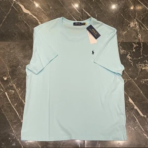 Ljusblå t-shirt från Polo Ralph Lauren - Snygg ljusblå t-shirt från Polo Ralph Lauren med klassisk rund hals och korta ärmar. T-shirten har den ikoniska mörkblå logobroderingen på bröstet och är gjord i mjuk bomull för en skön känsla. Perfekt till jeans eller shorts. Passar L/XL