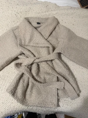 Beige teddyjacka med knytskärp - Supermjuk och fluffig teddyjacka i beige med bred krage och knytskärp i midjan. Jackan har en avslappnad passform och är tillverkad i ett mysigt teddy-material som håller dig varm. Perfekt för kyliga dagar när du vill vara både bekväm och snygg.