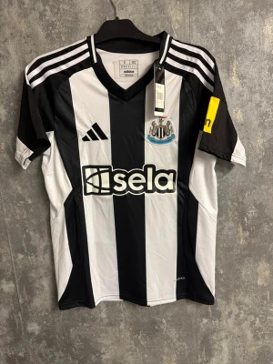 Newcastle Isak - Ny Small - Säljer en svartvit randig Newcastle United matchtröja från Adidas med klubbmärke, sponsortryck och nummer 14 ISAK på ryggen. Tröjan har klassiska Adidas-ränder på axlarna och gul detalj på ärmen. Perfekt för dig som älskar fotboll och Premier League.