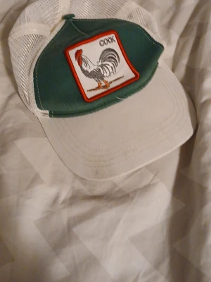 Grön och vit truckerkeps med tupp - Säljer en grön och vit truckerkeps med mesh baktill och justerbart plastspänne. Framtill sitter ett patch med en tupp och texten 'COCK'. Perfekt för dig som vill sticka ut med en cool och retro keps.