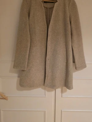 Beige kappa med rak passform - Stilren beige kappa med rak passform och diskreta fickor i sidorna. Kappan har dold knäppning framtill. Använd fåtal gånger