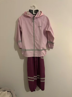 Rosa regnställ från FUNK - Säljer ett tvådelat regnställ från FUNK med ljusrosa jacka och mörkrosa byxor. Jackan har huva, dragkedja och reflexdetaljer på ärmar och nederkant. Byxorna har resår i benslut och reflexband. Perfekt för regniga dagar!