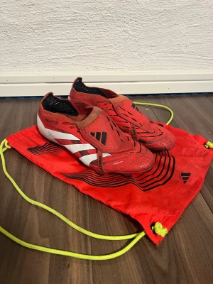 Adidas Predator röda fotbollsskor - Säljer ett par röda Adidas Predator fotbollsskor med vita och svarta detaljer. Skorna har en strömlinjeformad design utan snörning, med texten 'PREDATOR' under sulan och levereras med en matchande röd påse. Perfekta för dig som vill sticka ut på planen.