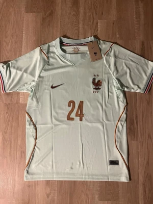 Frankrike VM-2026 Fotbollströja #24 Cherki - Ljusgrön fotbollströja för Frankrikes landslag med bronsfärgade detaljer, nummer 24 och namnet Cherki på ryggen. Tröjan har korta ärmar, broderad tupp och FFF-logga samt randiga muddar i blått, vitt och rött. Tillverkad i lätt funktionsmaterial. HELT NY!