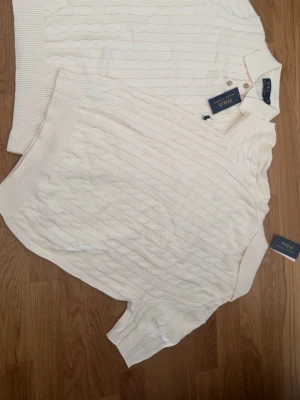 Vit kabelstickad pikétröja Polo Ralph Lauren - Kabelstickad pikétröja i krämvit färg från Polo Ralph Lauren. Tröjan har korta ärmar, ribbade muddar och klassisk krage med knappar. Perfekt för en stilren och avslappnad look. Materialet känns mjukt och exklusivt.