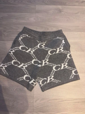 Grå stickade shorts med CB-mönster - Snygga grå stickade shorts med vitt CB-mönster över hela plagget. Mjukt material och bred resår i midjan för skön passform. Perfekta för en avslappnad och trendig look.