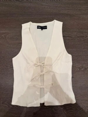 Vit väst med snörning från Zara - Stilren vit väst från Zara med v-ringad hals och snygg snörning framtill. Västen har en enkel, rak passform och är tillverkad i ett mjukt tyg som ger en clean look. Perfekt att styla över en t-shirt eller skjorta för en trendig outfit.