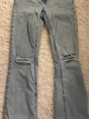Ljusblå slitna bootcut jeans - Säljer ett par ljusblå bootcut jeans med slitningar över knäna och cool röd zigzag-detalj på bakfickan. Jeansen har klassisk femficksdesign och är tillverkade i denim. Perfekta för en avslappnad och trendig look.