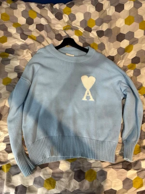 Ami Paris tröja - Ljusblå stickad tröja med rund hals och ribbade muddar. På bröstet finns en vit hjärtdetalj med bokstaven 'A' under. Tröjan har en avslappnad passform och är perfekt för en chill och stilren look.