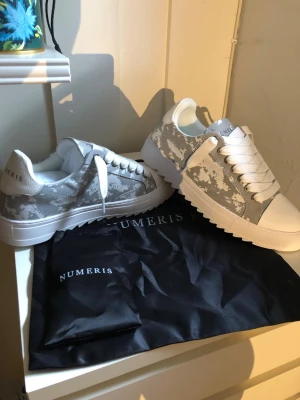 Numeris vita och grå camo sneakers - Snygga sneakers från Numeris i vitt och grått med camo-mönster. Skorna har vita snören, grov sula och detaljer i läderimitation. Perfekta för dig som vill sticka ut med en modern och fräsch look.