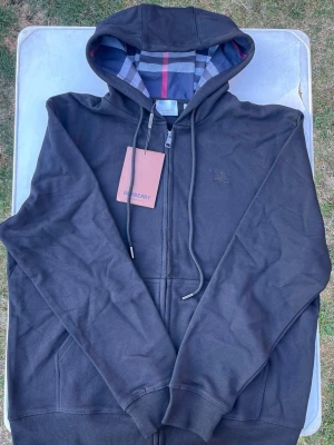 Svart hoodie från Burberry - Snygg svart hoodie från Burberry med dragkedja och klassiskt rutigt foder i huvan. Broderad Burberry-logga på bröstet och fickor framtill. Tillverkad i mjukt bomullsmaterial och har justerbara snören i huvan. Perfekt för en stilren och avslappnad look.