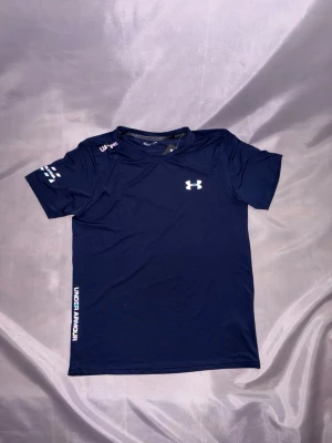 Under Armour tränings t-shirt  - Snyggt träningsset från Under Armour med en marinblå t-shirt och svarta shorts. Båda plaggen har logga och text i vitt. Tillverkade i ett lätt och snabbtorkande material som passar perfekt till gymmet eller löpning.