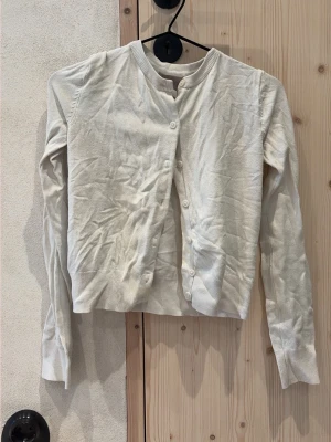 Vit basic kofta med rund hals - En enkel vit kofta med rund halsringning och knäppning framtill. Långärmad modell med ribbade muddar vid ärmslut och nederkant. Perfekt att slänga över en topp för en clean look.