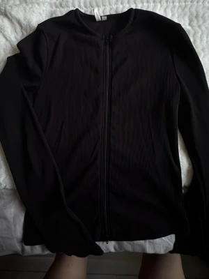 Svart ribbad ziptröja - Säljer en svart ribbad ziptröja med lång ärm och rund hals. Tröjan har dragkedja framtill och är tillverkad i ett stretchigt material som ger en snygg passform. Perfekt att slänga på sig över en t-shirt eller linne.