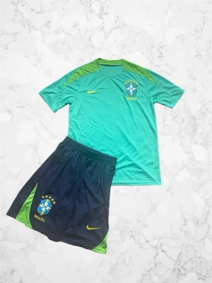 Brasilien Nike fotbollskit grön/blå - Snyggt Brasilien fotbollskit från Nike med grön t-shirt och blå shorts. Tröjan har gula detaljer och Brasilien-emblem på bröstet, medan shortsen har samma emblem och gula Nike-logga. Perfekt för träning eller match. Tillverkad i lätt och ventilerande material.