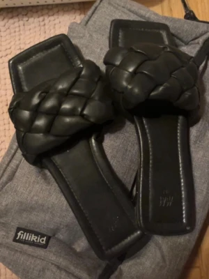 Svarta flätade sandaler från H&M - Stilrena svarta sandaler från H&M med breda, flätade remmar i vadderat konstläder. De har fyrkantig tå och platt sula, vilket ger en trendig look. Perfekta för dig som gillar minimalistisk och modern stil.