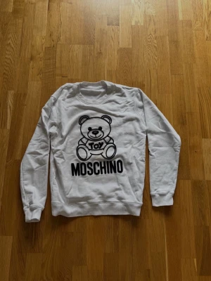Vit Moschino sweatshirt med björn - Vit sweatshirt från Moschino med stort svart tryck av en söt björn och texten 'Toy Moschino' på framsidan. Tröjan har rund halsringning, ribbade muddar och är tillverkad i mjukt material. Perfekt för en trendig och avslappnad look.