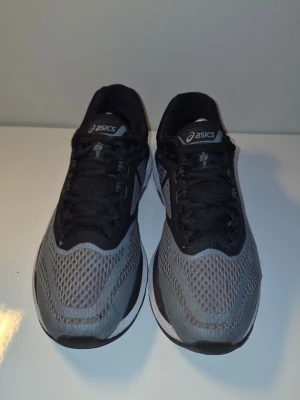 Gråa Asics sneakers med mesh - Snygga sneakers från Asics i grått och svart med ventilerande mesh-ovandel och klassisk logga på sidan. Skorna har svarta snören och en vit sula med svarta detaljer. Perfekta för dig som gillar sportig stil och komfort.