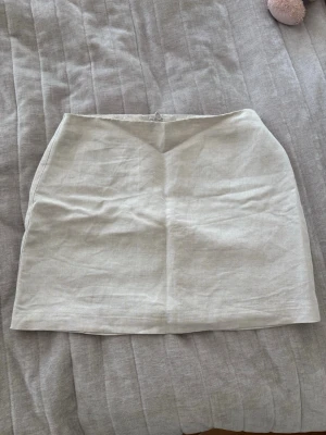 Beige linneshorts kjol från H&M - Säljer en stilren beige kortkjol från H&M i linne. Kjolen har en enkel, rak passform och dragkedja bak. Perfekt för dig som gillar minimalistisk och fräsch stil. Passar till både sneakers och sandaler.