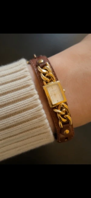 Michael Kors läderklocka med gulddetaljer - Stilren klocka med rektangulär urtavla i guld och armband i brunt läder. Klockan har snygga guldfärgade kedjedetaljer på bandet och klassiskt spänne. Perfekt accessoar för att lyfta din outfit. OBS!Behövs lämnas in till klockmästare!