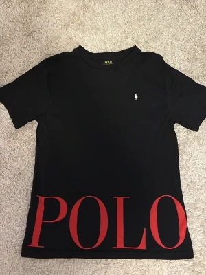 Svart POLO t-shirt med röd logga - Svart t-shirt från Polo Ralph Lauren med stor röd POLO-logga längst ner och klassisk liten vit logga på bröstet. T-shirten har rund hals och korta ärmar, tillverkad i mjuk bomull för en skön känsla.