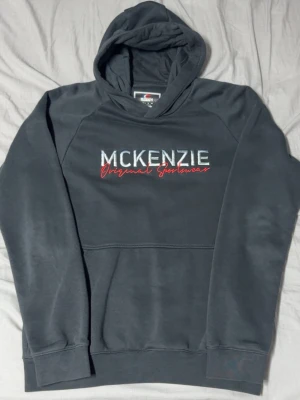 Svart hoodie från McKenzie - Snygg svart hoodie från McKenzie med broderad text 'MCKENZIE Original Sportswear' i vitt och rött på bröstet. Klassisk känguruficka framtill och huva. Perfekt för en avslappnad och sportig stil. Sista bilden visar hur den sitter fast på bilden är det bara en annan färg men exakt samma tröjmodell.