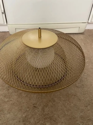 Guldig lampskärm i metallnät - Säljer en rund lampskärm i guldigt metallnät med modern design. Skärmen har ett nätmönster och en cylinderformad del i mitten för lampfäste. Passar perfekt för dig som vill ha en unik och stilren belysning i rummet.
