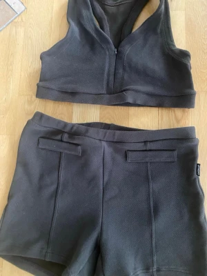 Mullvard färgad träningsset från Casall,  - Säljer ett sportigt tvådelat set från Casall i mörkgrå färg. Setet består av en croppad topp med dragkedja framtill och ett par matchande shorts med fickdetaljer bak. Materialet är mjukt och stretchigt, perfekt för en aktiv stil.
