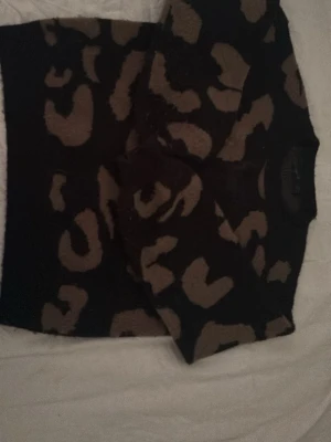 Svart stickad tröja med leopardmönster - Cool svart stickad tröja med stora beige leopardfläckar. Tröjan har rund hals och långa ärmar. Perfekt för dig som vill sticka ut med ett djurmönstrat plagg i garderoben. Materialet känns mjukt och bekvämt.