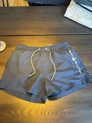 Marinblå badbyxor Sail Racing - Snygga marinblå badbyxor från Sail Racing med vitt snöre i midjan och logga tryckt på ena benet. Klassisk kort modell i lätt och snabbtorkande material, perfekta för stranden eller poolen. Pris kan diskuteras!