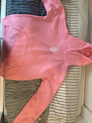 Rosa hoodie från Self Love Club - Säljer en rosa hoodie från Self Love Club med broderad logga och text på bröstet samt ärmen. Hoodien har en stor magficka och klassisk huva. Tillverkad i mjukt material som känns skönt mot huden. Perfekt för dig som gillar streetwear och vill ha en poppig färg i garderoben.