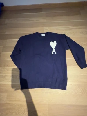Mörkblå stickad tröja från Ami - Mörkblå stickad tröja från Ami med rund halsringning och ribbade muddar. På bröstet finns ett vitt hjärta och bokstaven A. Tröjan har en klassisk passform och är perfekt för dig som gillar stilrena detaljer.